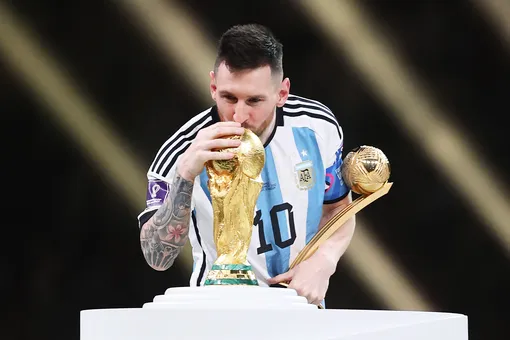 Лионель Месси целует Кубок победителей Чемпионата мира по футболу 2022 года, держа в руках награду «Золотая бутса» от adidas